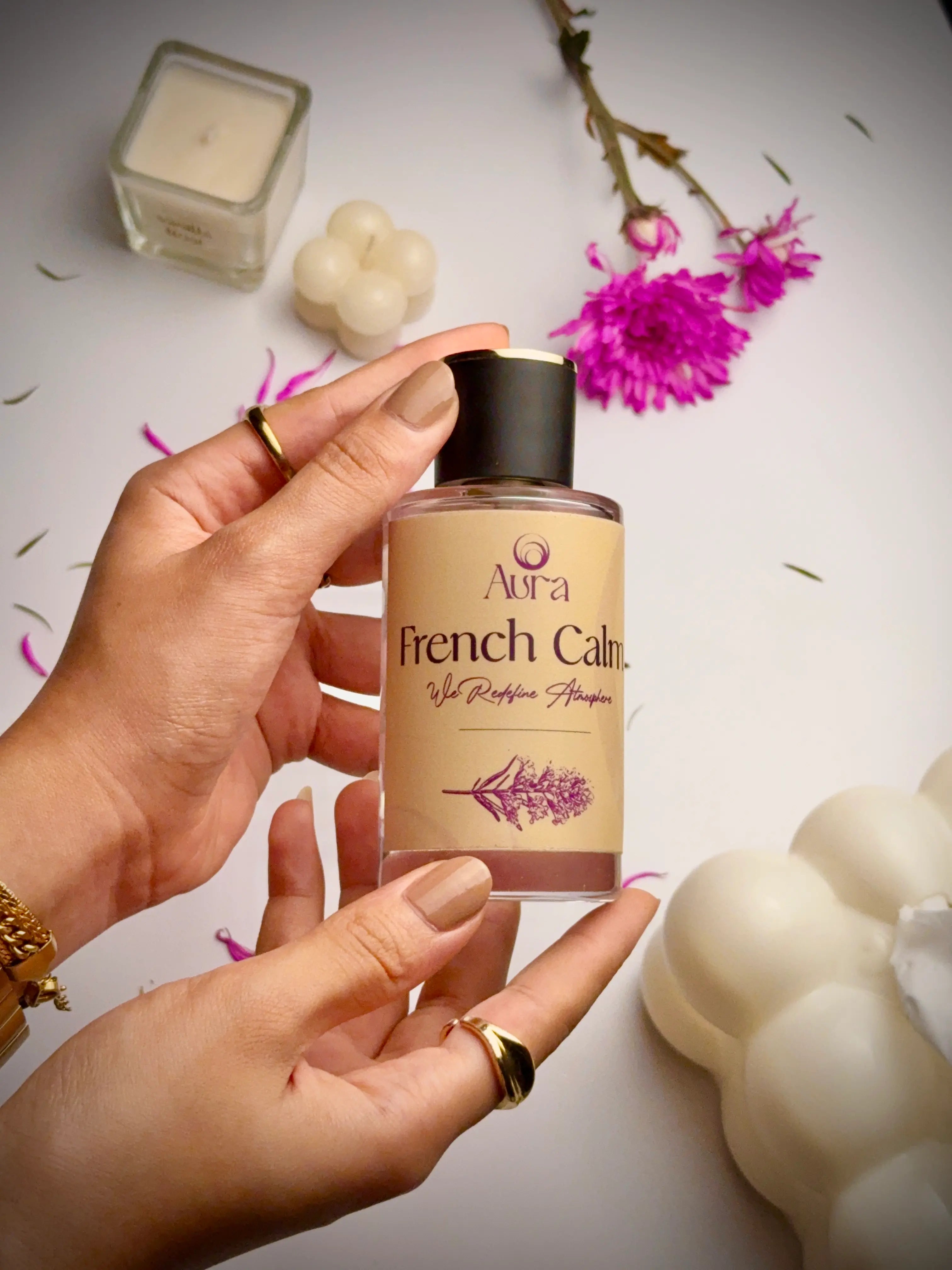 Hand holding a bottle of 'Aura French Calm' product with flowers and candles in the background