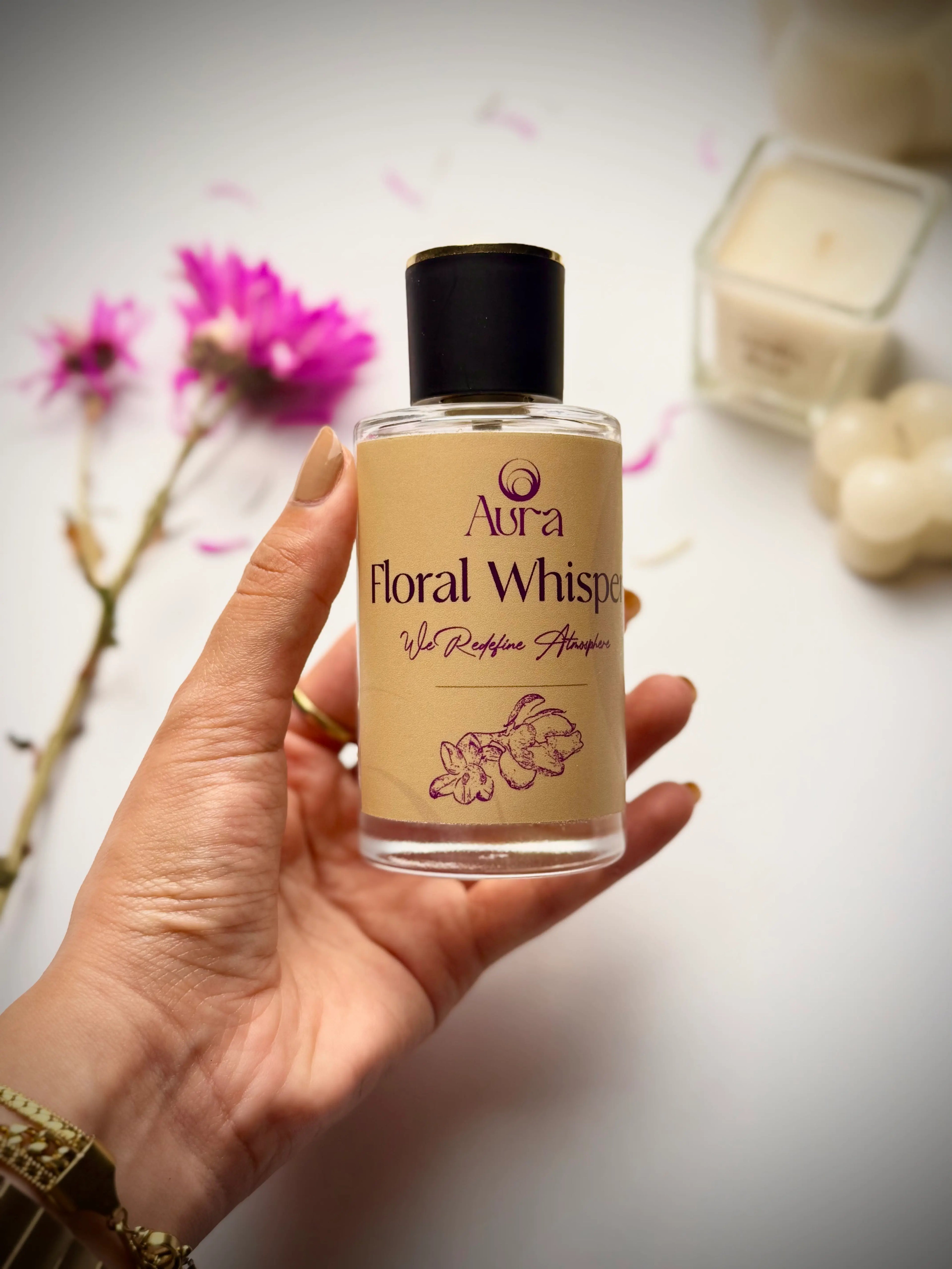Hand holding a bottle of 'Aura Floral Whisp' perfume with flowers and a box in the background.