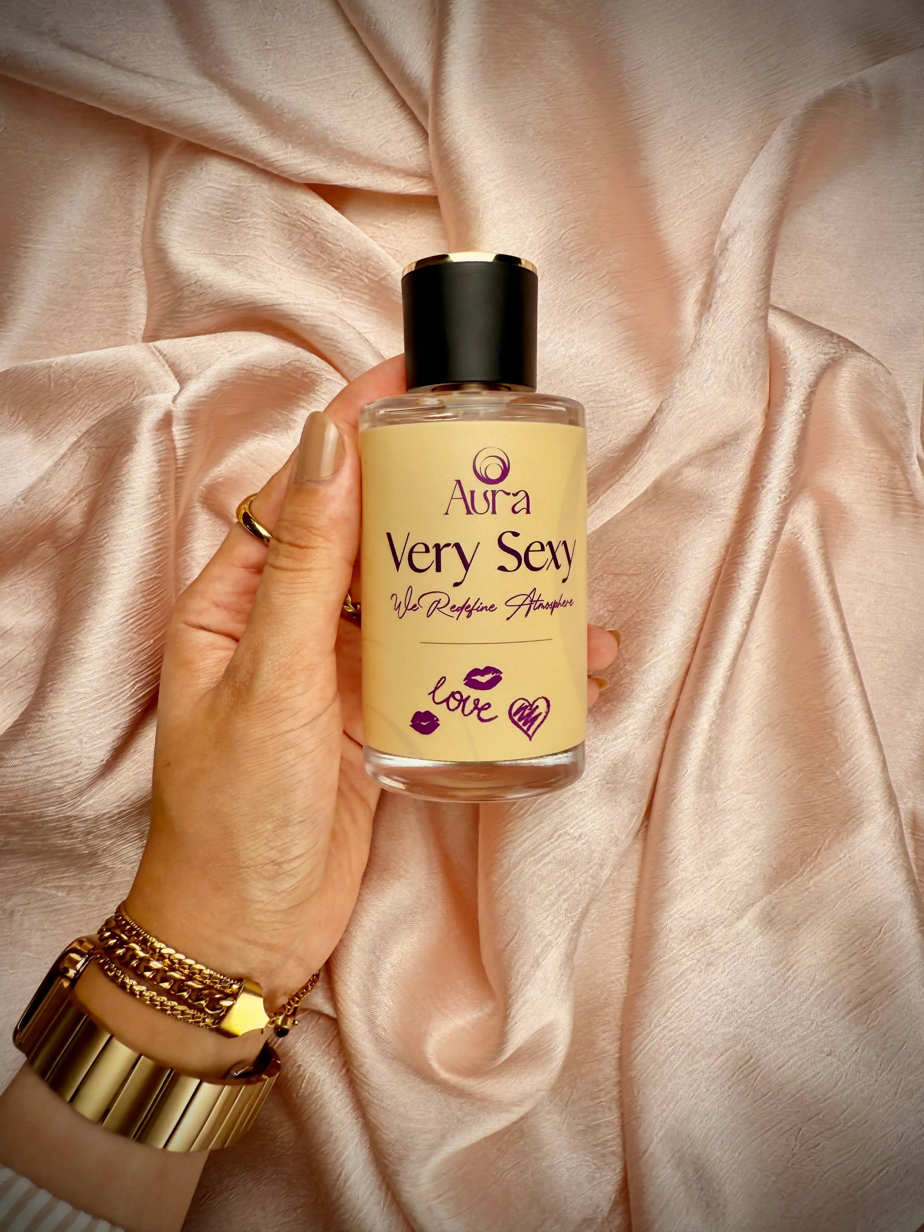 Hand holding a bottle labeled 'Aura Very Sexy' against a soft pink fabric background