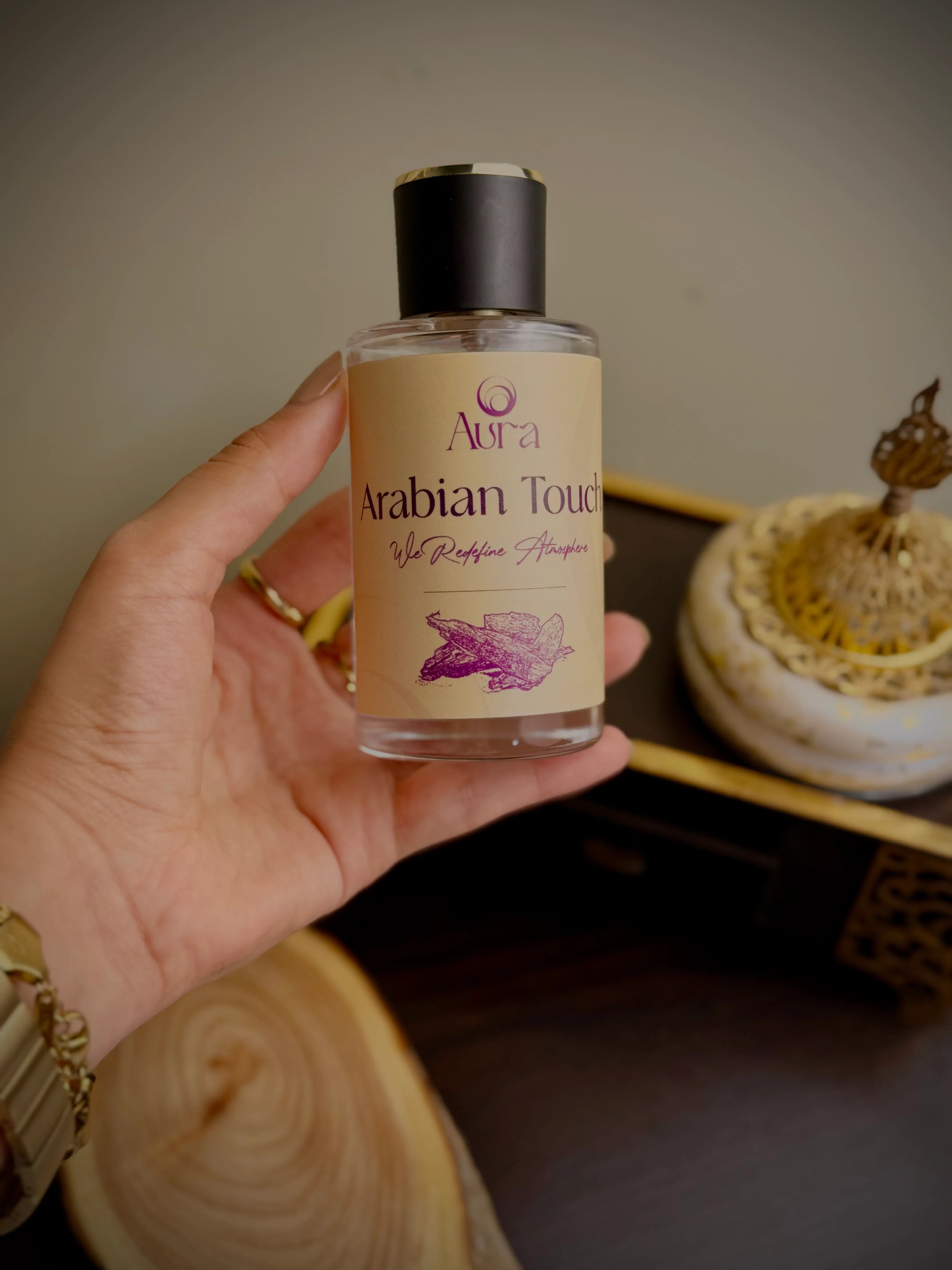 Hand holding a bottle of 'Aura Arabian Touch' product with a blurred background
